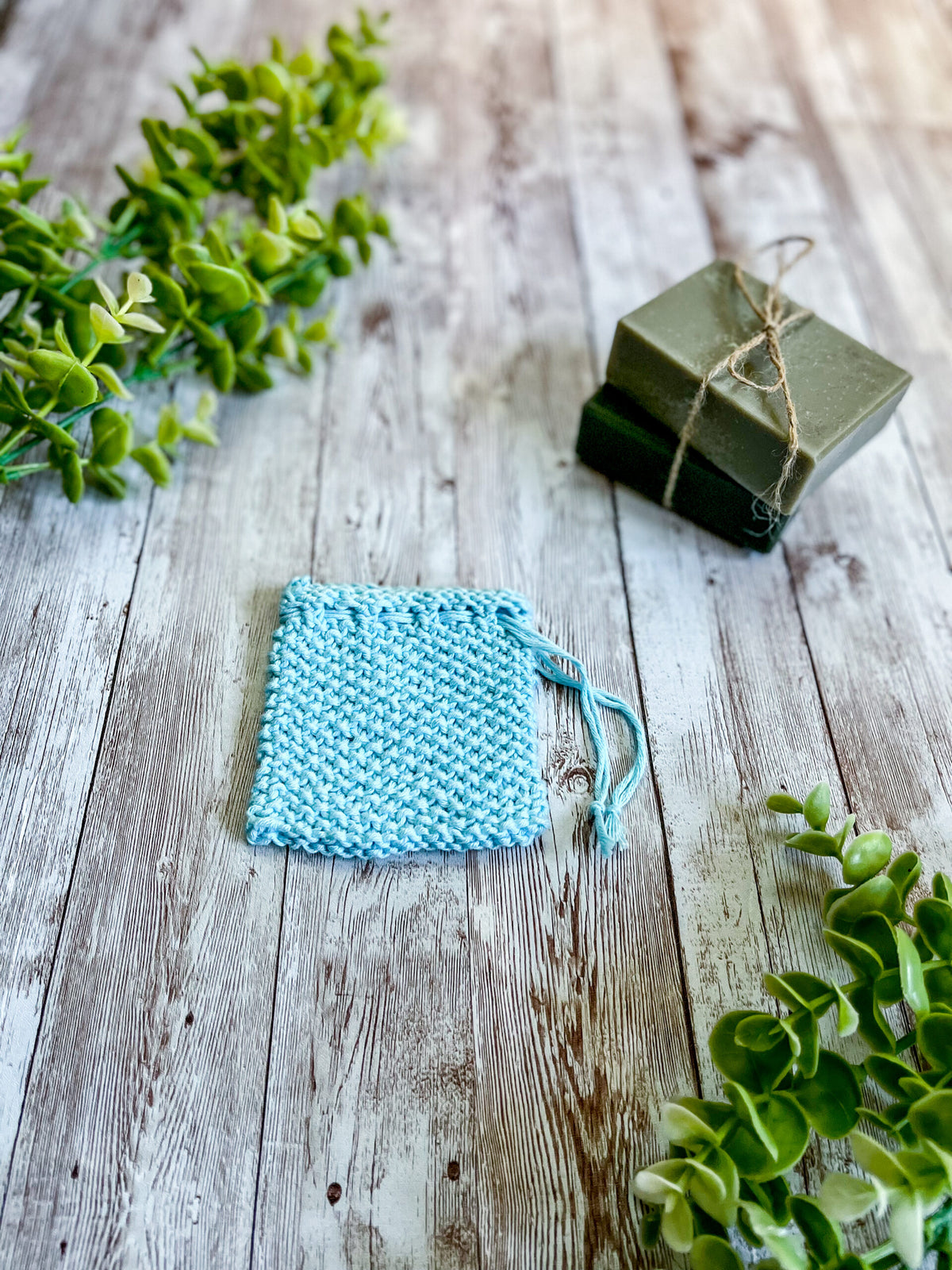 A blue bamboo/cotton soap saver rests on a wood plank next to 2 bars of soap wrapped with twine and a sprig of eucalyptus leaves