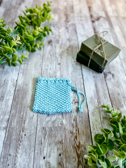 A blue bamboo/cotton soap saver rests on a wood plank next to 2 bars of soap wrapped with twine and a sprig of eucalyptus leaves