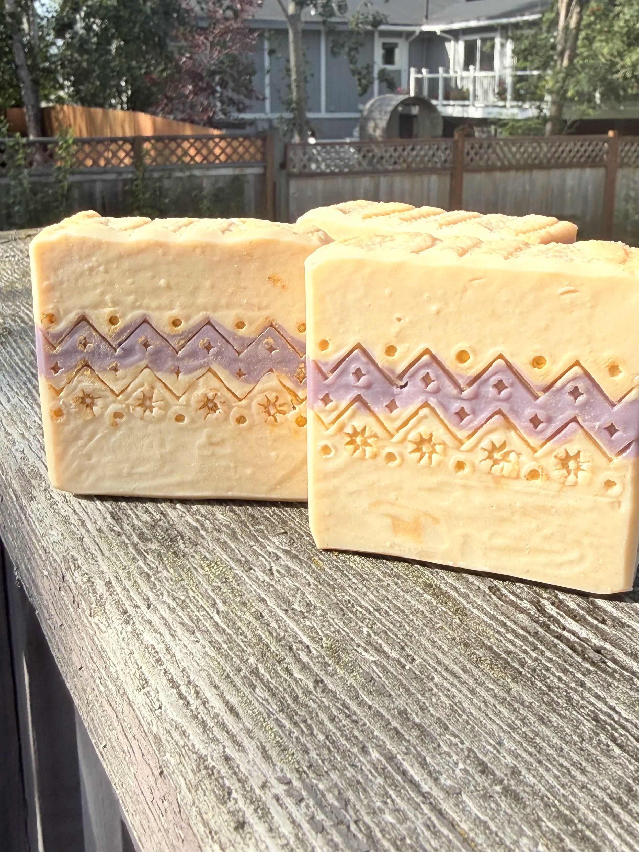Two handcrafted Ugly Sweater holiday soap bars with cream and lavender sweater-inspired patterns, displayed on rustic wood outdoors.