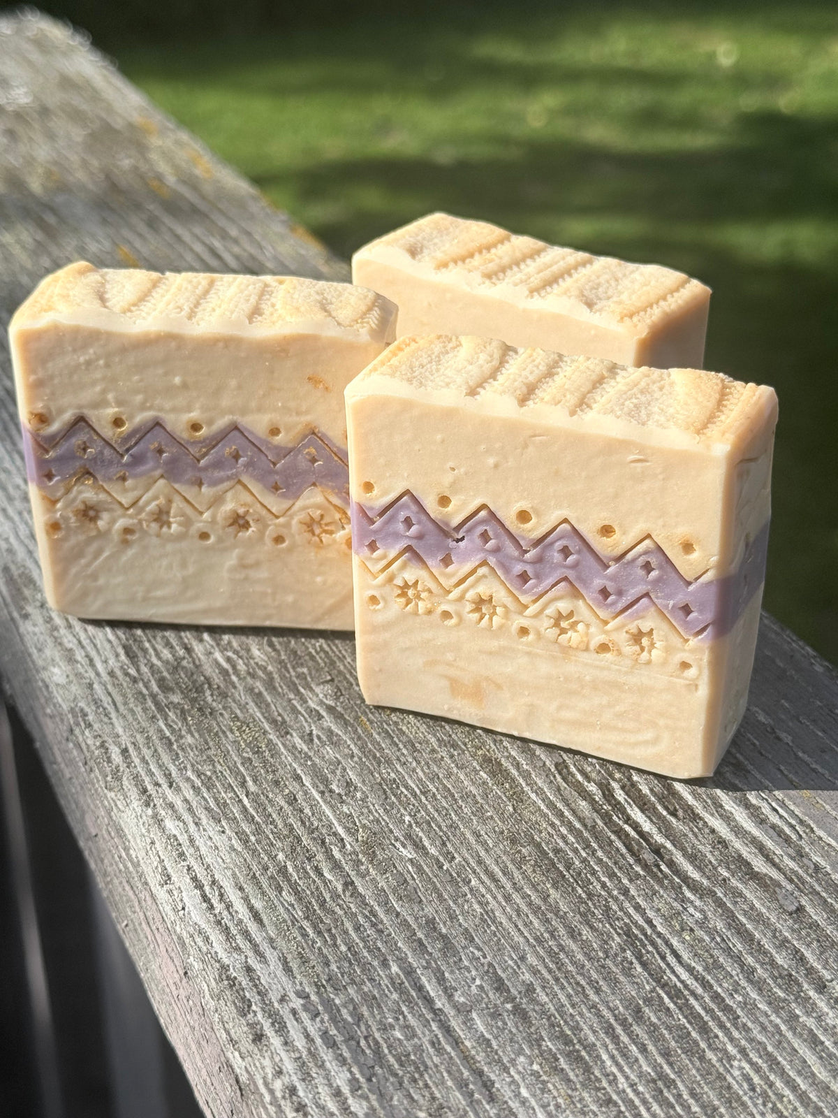 Three handcrafted Ugly Sweater holiday soap bars with cream and lavender sweater-inspired patterns, displayed on rustic wood outdoors.
