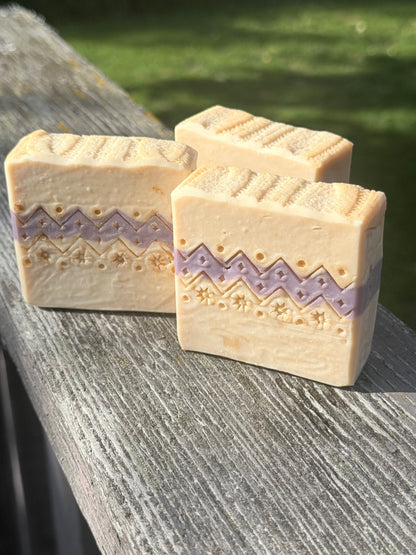 Three handcrafted Ugly Sweater holiday soap bars with cream and lavender sweater-inspired patterns, displayed on rustic wood outdoors.