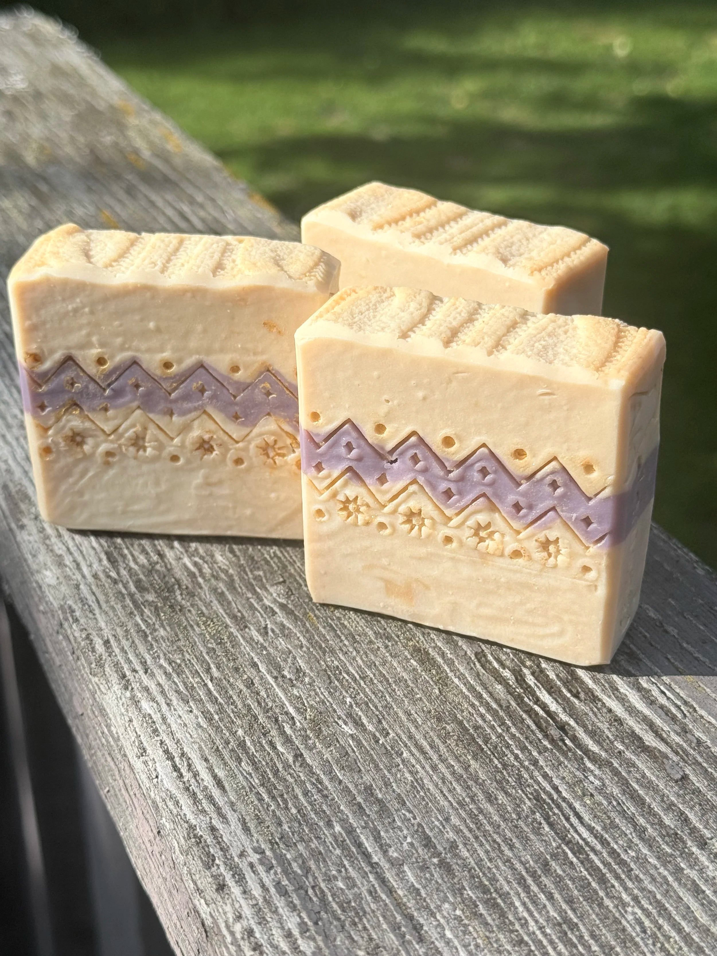 Three handcrafted Ugly Sweater holiday soap bars with cream and lavender sweater-inspired patterns, displayed on rustic wood outdoors.