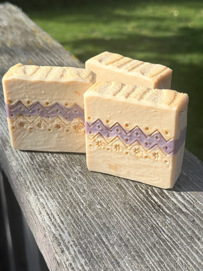 Three handcrafted Ugly Sweater holiday soap bars with cream and lavender sweater-inspired patterns, displayed on rustic wood outdoors.
