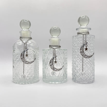 Three clear glass perfume bottles with stoppers and silver crescent moon and star chain ornaments, displayed against a light gray background.
