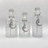 Three clear glass perfume bottles with stoppers and silver crescent moon and star chain ornaments, displayed against a light gray background.
