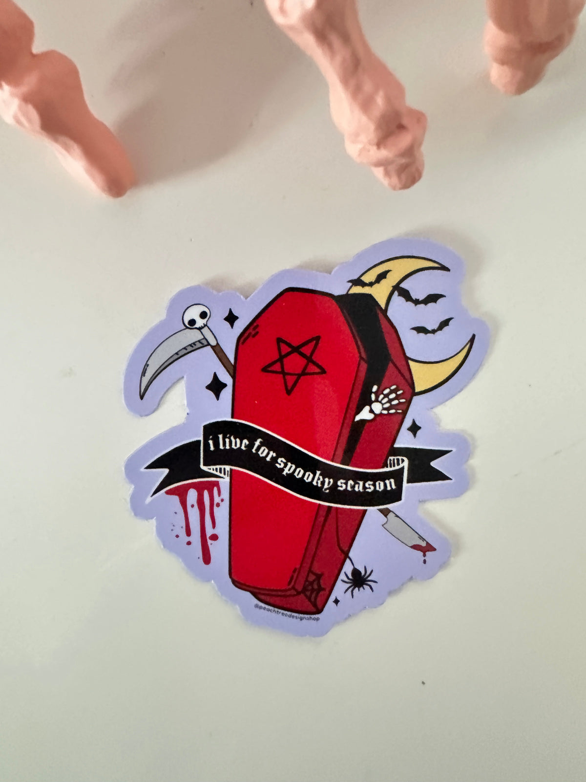 A die-cut sticker featuring a red coffin with a black band, black pentagram, white skeleton hand, crescent moon, scythe, and a black banner reading &