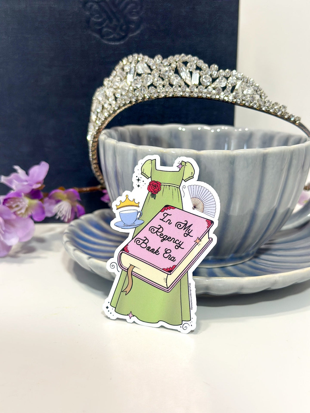 A die-cut sticker featuring an illustration of a green dress, pink book THAT SAYS "IN MY REGENCY ERA" in script, and teacup, placed in front of a gray ceramic teacup and saucer with a silver tiara on a white surface.