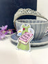 A die-cut sticker featuring an illustration of a green dress, pink book THAT SAYS "IN MY REGENCY ERA" in script, and teacup, placed in front of a gray ceramic teacup and saucer with a silver tiara on a white surface.