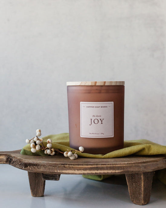 Joy candle in amber glass jar on dark wood surface, evoking floral and citrus notes – Fig &amp; Grove