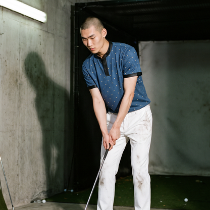 A man at an indoor golf driving range wearing a blue patterned polo shirt and white pants, preparing to hit a golf ball with a driver. Golf clubs are stored in the background, and a golf ball monitor is visible on the ground.