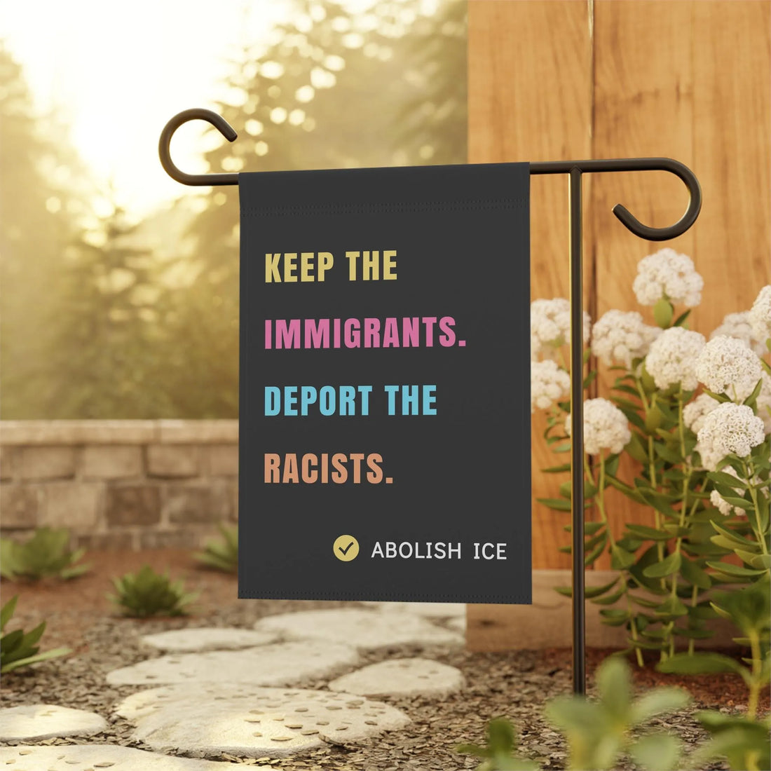 Black garden flag with bold, multicolored text reading &amp;quot;KEEP THE IMMIGRANTS. DEPORT THE RACISTS. ABOLISH ICE,&amp;quot; displayed outdoors on a metal flag stand surrounded by flowers and greenery. Pro-immigrant, anti-racist activist flag for yard or garden decor.