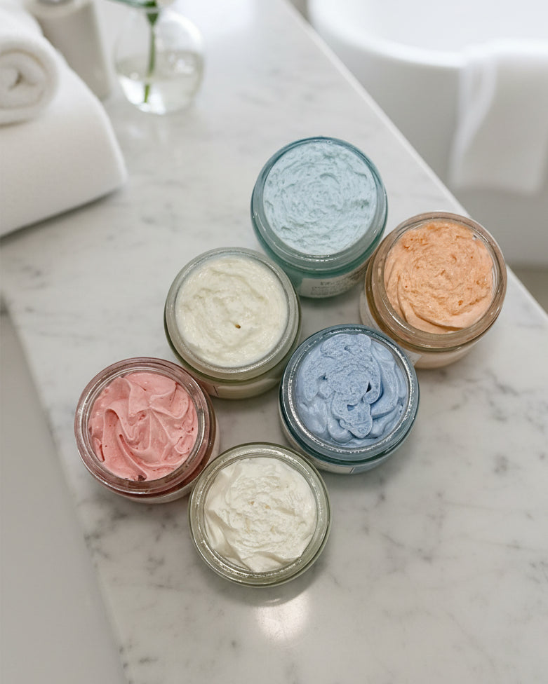 Six small jars with colorful whipped body butter on a white surface