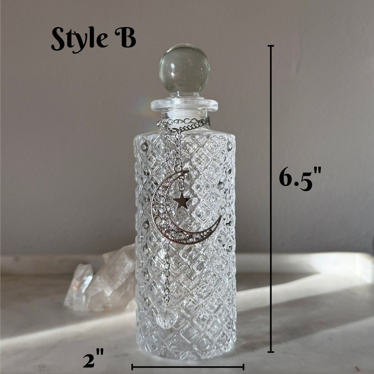 A clear textured glass bottle with a round stopper and a silver crescent moon and star pendant, shown with dimension labels reading 6.5 inches tall and 2 inches wide on a neutral background.