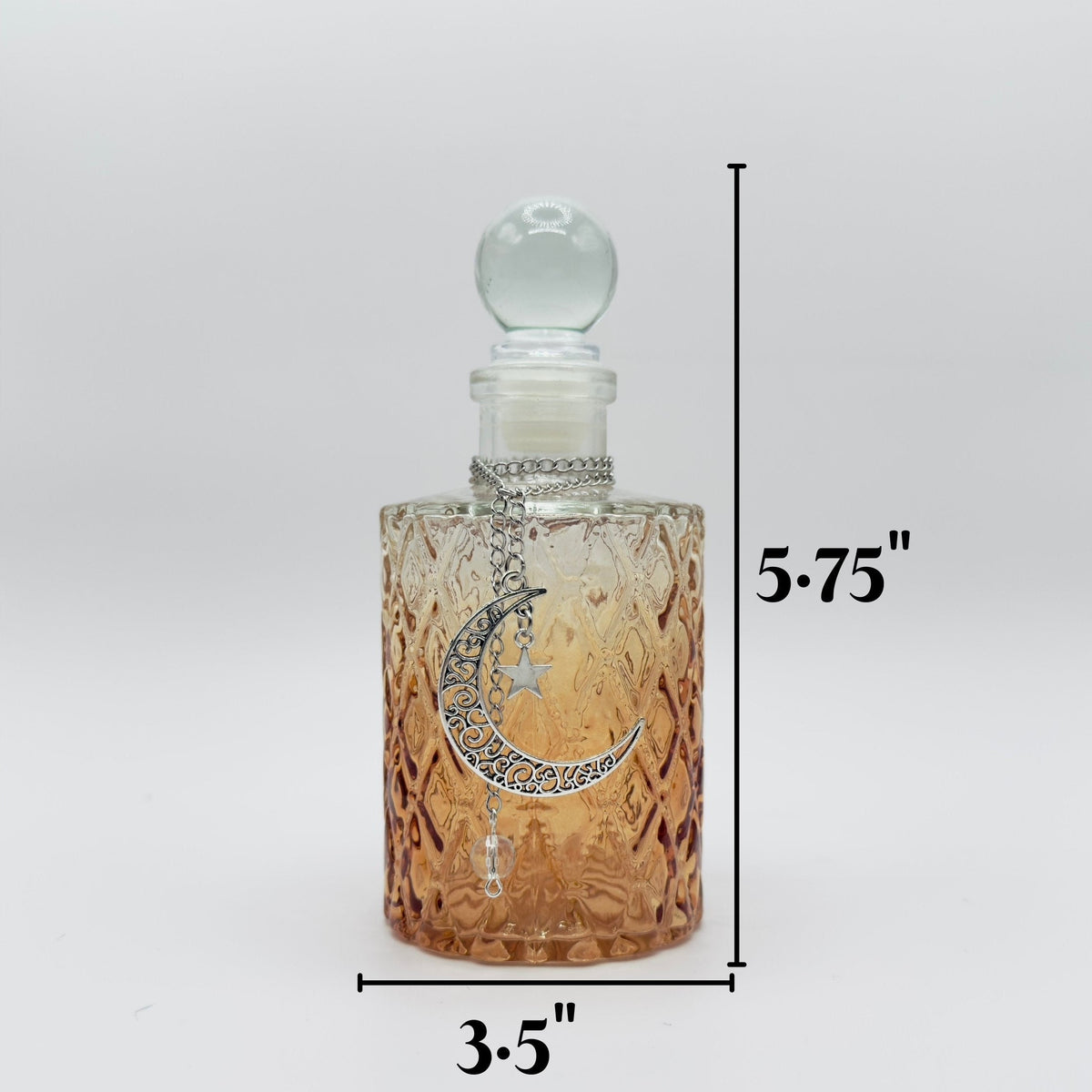 A clear glass perfume bottle with a textured amber-tinted body, a silver crescent moon and star pendant, and a clear glass stopper. Black dimension lines indicate a height of 5.