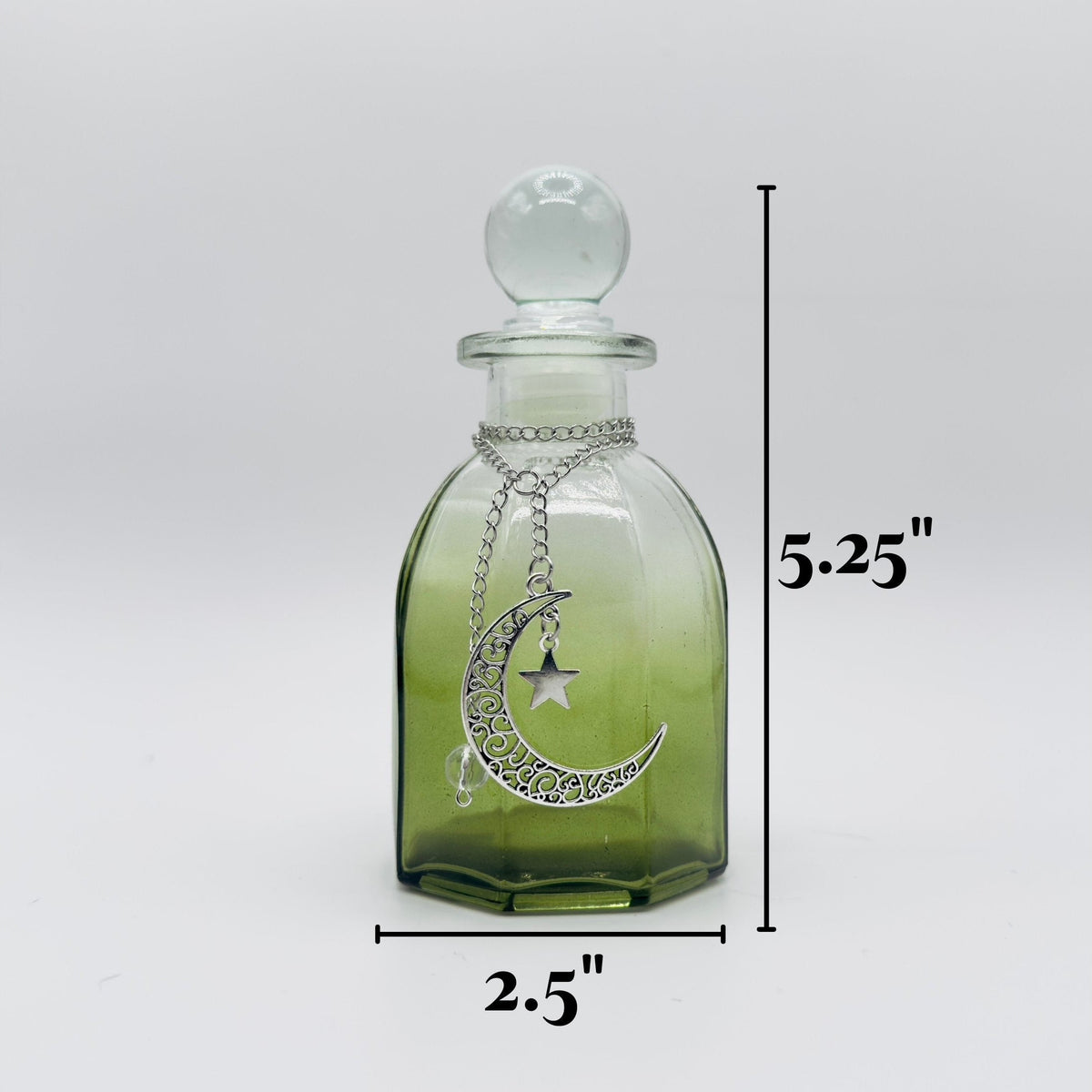 A small clear glass bottle with a rounded stopper and a silver crescent moon and star pendant, shown on a light gray background with dimension lines indicating 5.25 inches tall and 2.