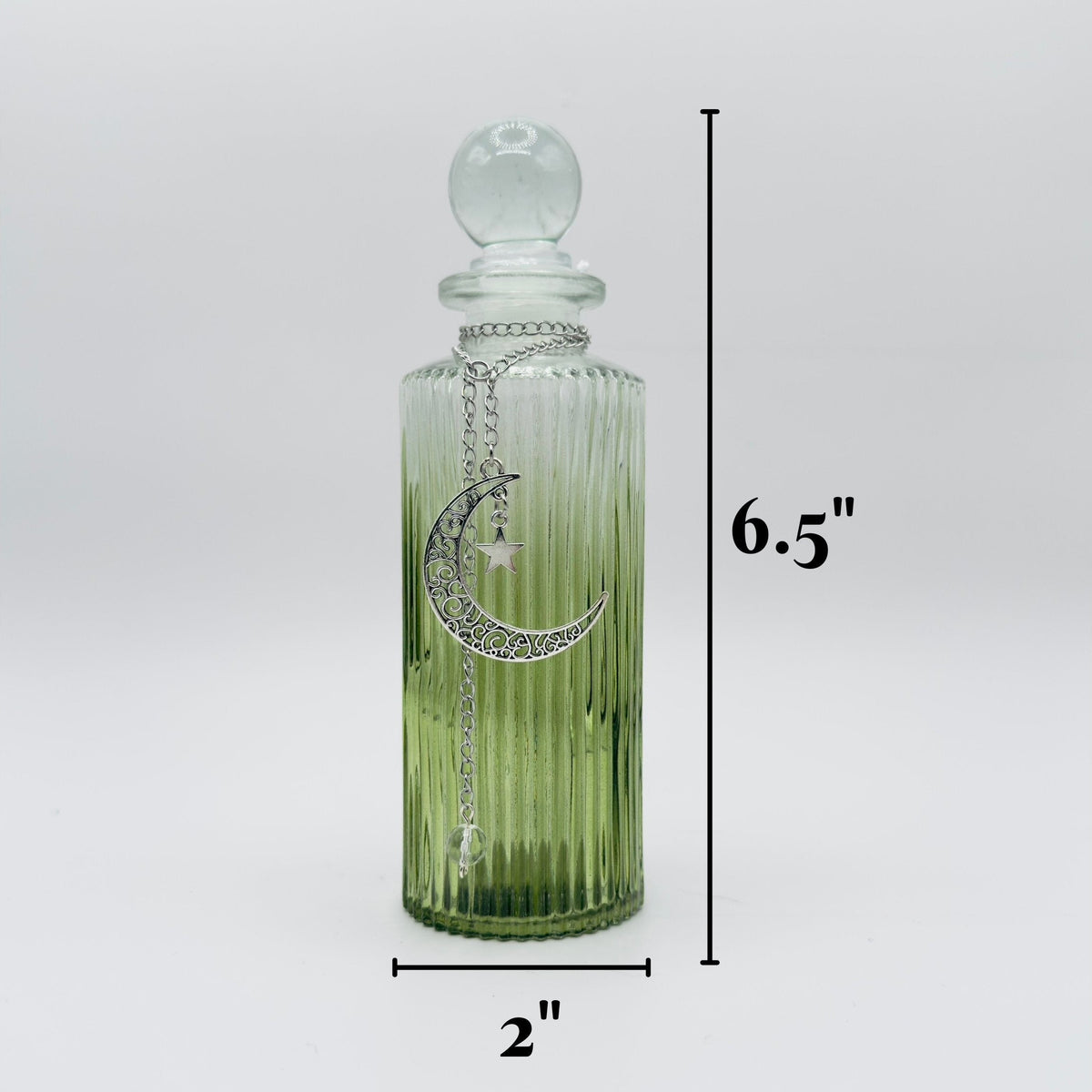 A ribbed clear glass bottle with a matching round stopper, featuring a silver crescent moon charm with rhinestones and a small white star. The bottle has a green-to-clear gradient tint.