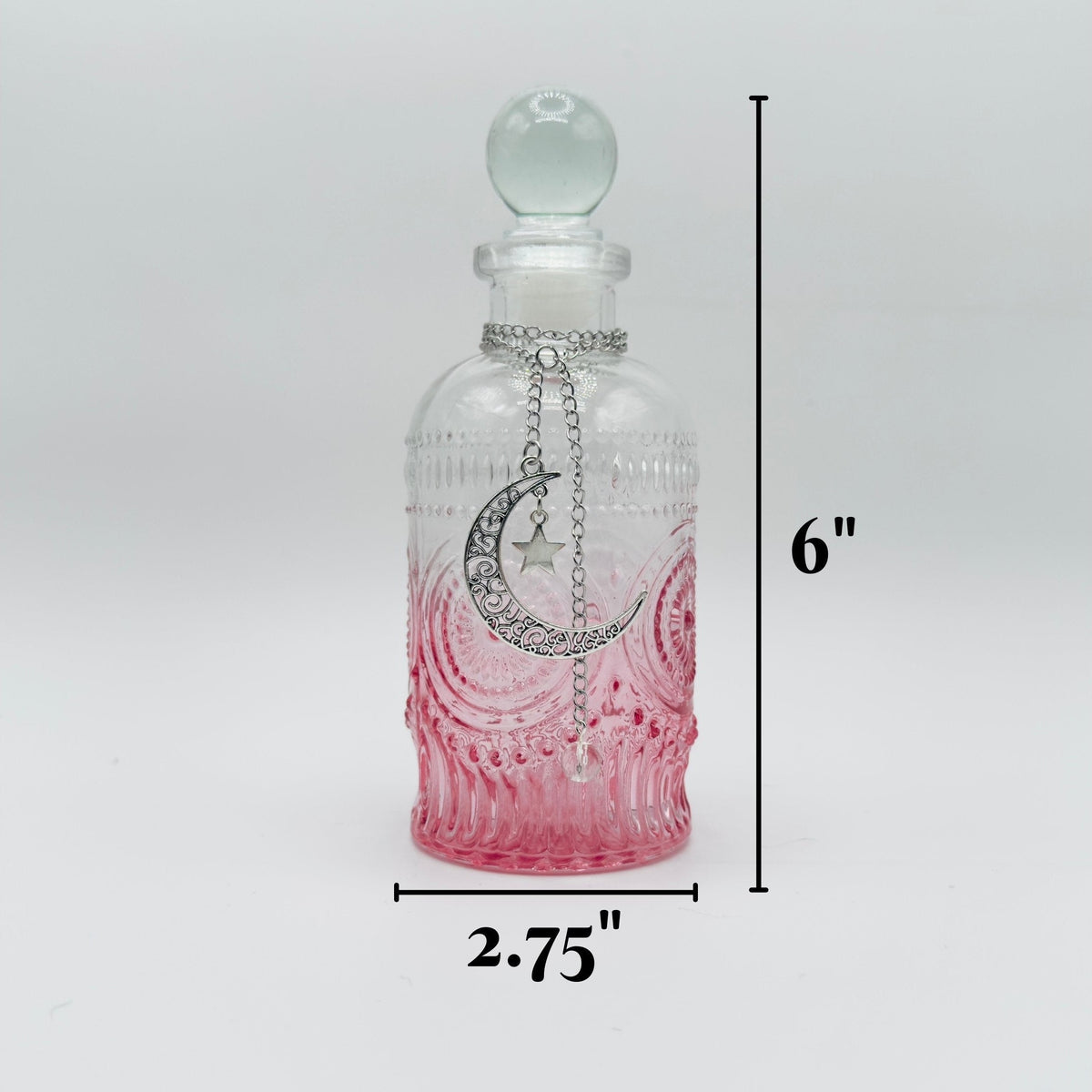 A small clear glass bottle with a rounded stopper, decorated with a silver crescent moon and star pendant, shown with black dimension lines indicating a height of 6 inches and a width of 2.75 inches on a light gray background.
