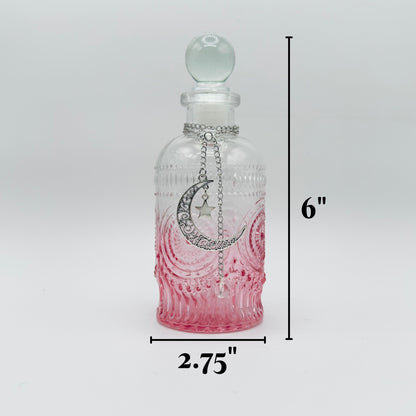 A small clear glass bottle with a rounded stopper, decorated with a silver crescent moon and star pendant, shown with black dimension lines indicating a height of 6 inches and a width of 2.75 inches on a light gray background.