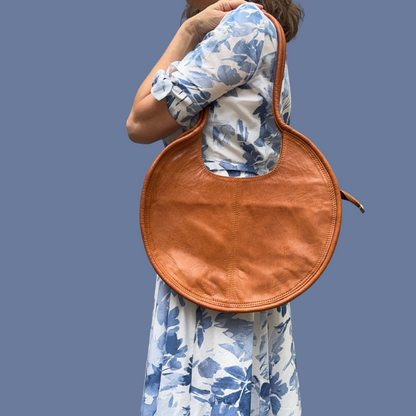 A person wearing a floral dress is seen from the back, carrying an natural leather circular purse over their shoulder.