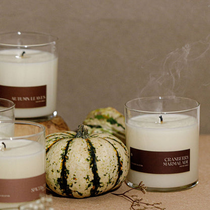 A styled arrangement of several white scented candles in clear glass jars with brown labels, set on a tabletop with small gourds and a neutral wall background. Visible label text includes &