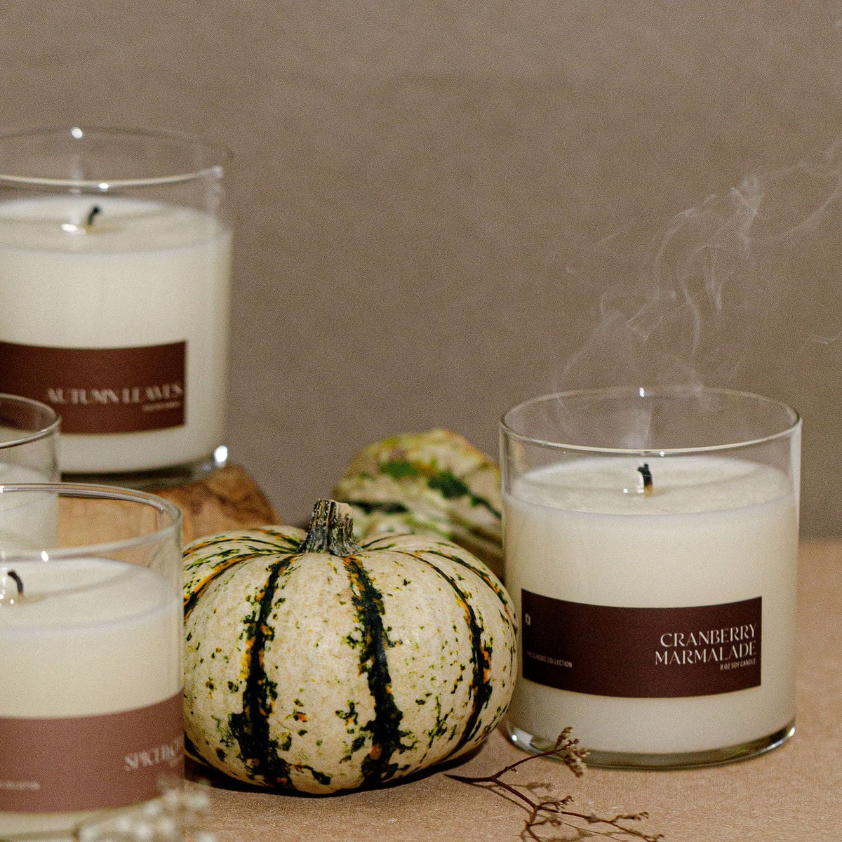 A styled arrangement of several white scented candles in clear glass jars with brown labels, set on a tabletop with small gourds and a neutral wall background. Visible label text includes &