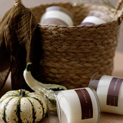 Two white glass jar candles with dark brown labels are styled in a rustic autumn scene with a woven basket, a small green pumpkin, and a brown fabric bag.
