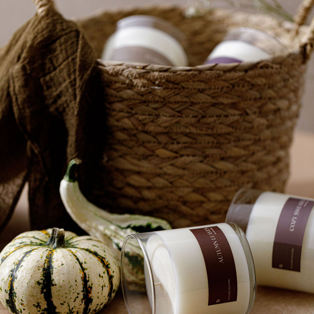 Two white glass jar candles with dark brown labels are styled in a rustic autumn scene with a woven basket, a small green pumpkin, and a brown fabric bag.