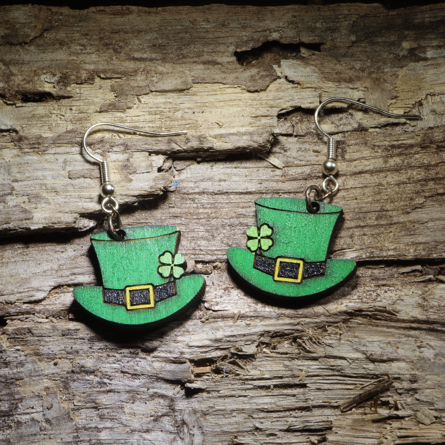 Pair of hand-painted green leprechaun hat wooden dangle earrings with shamrock, black band and gold buckle displayed on rustic wood