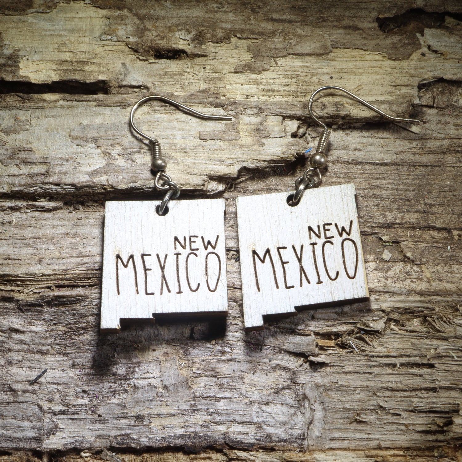 Pair of white New Mexico state-shaped wooden dangle earrings with New Mexico text displayed on rustic weathered wood