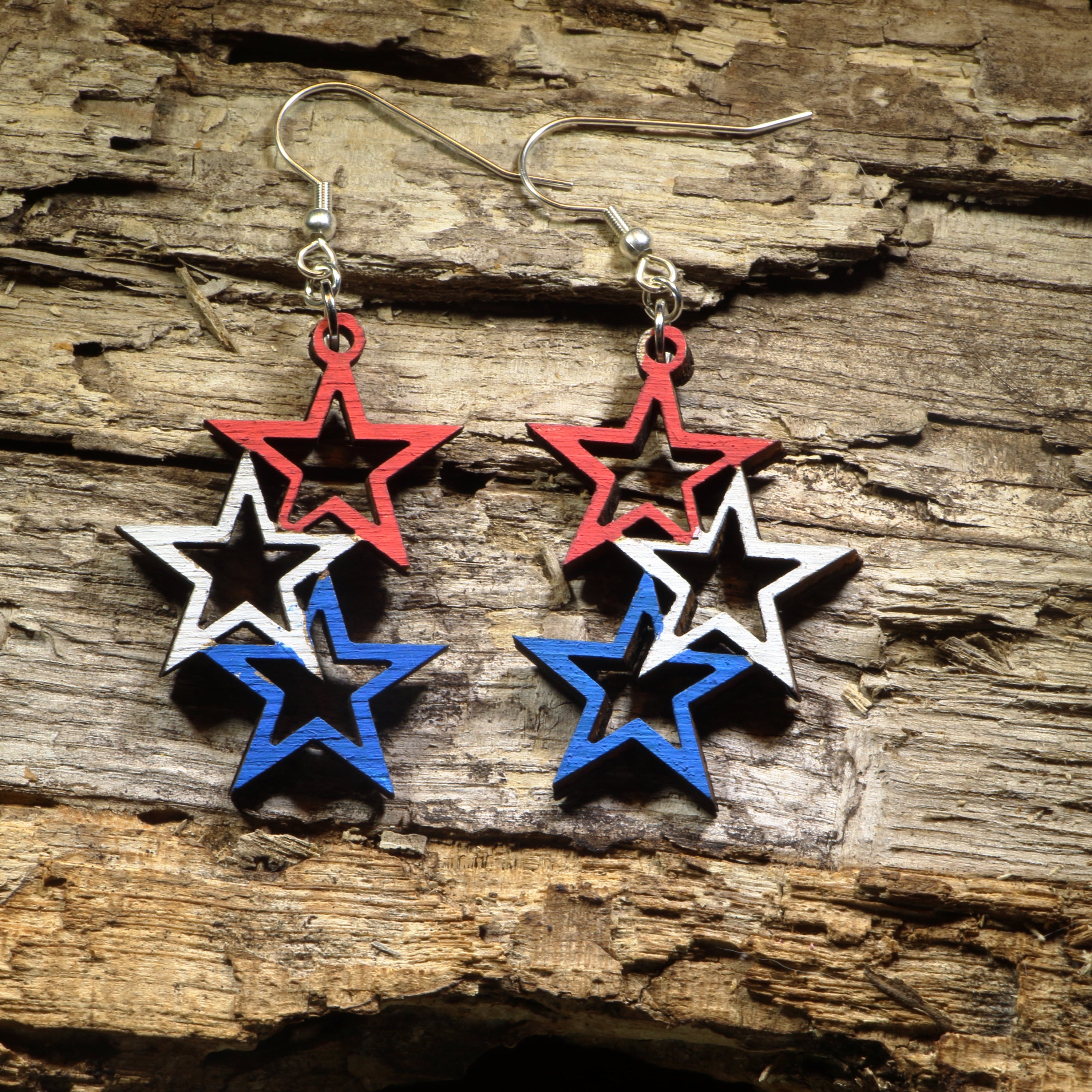 Hand-painted wooden patriotic star dangle earrings featuring layered red, white, and blue stars with a bold Americana design. Crafted from lightweight wood with hypoallergenic .925 sterling silver hooks, these festive earrings are perfect for Fourth of July celebrations, Independence Day, Memorial Day, Veterans Day, patriotic events, election season, Americana fashion, and anyone who loves showing their American pride.