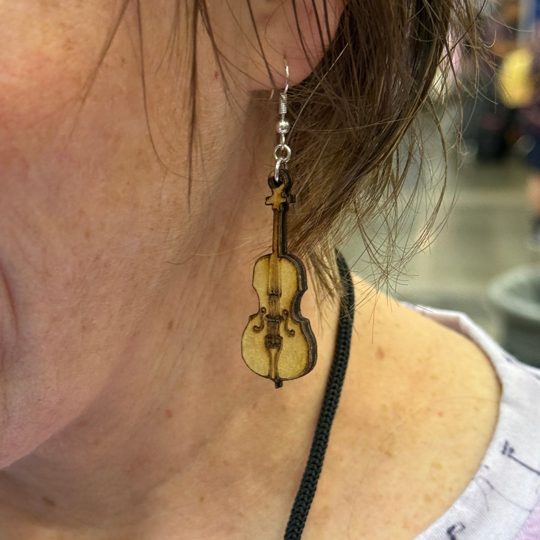 Person wearing a hand-painted wooden cello dangle earring with engraved strings and classic instrument details on a hypoallergenic .925 sterling silver hook, showing the lightweight size and elegant fit when worn by a cellist or classical music lover.