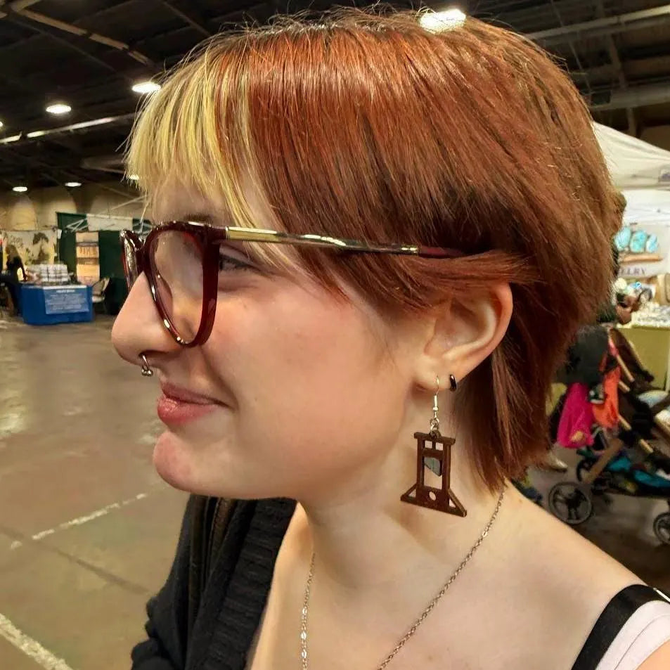 Person wearing hand-painted wooden guillotine dangle earrings inspired by the French Revolution, lightweight gothic history jewelry by Cate’s Concepts.