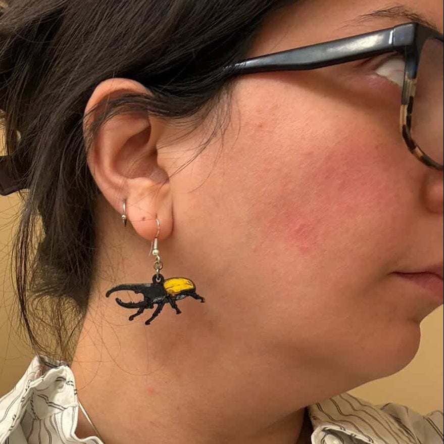 Person wearing hand-painted wooden Hercules beetle dangle earring with a black body and bright yellow wing covers on a hypoallergenic .925 sterling silver hook, showing the lightweight size and fit when worn.