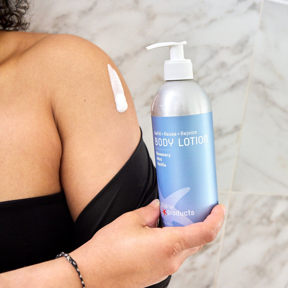 Person holding a plastic free bottle of rosemary mint vanilla eco friendly body lotion and lotion on her skin with a marble background