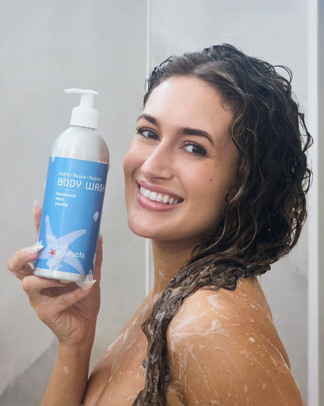 Woman holding an aluminum bottle of eco friendly rosemary mint vanilla body wash in a bathroom setting with lather.