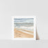 A framed art print featuring a watercolor painting of a coastline scene with sand, sea, and sky.