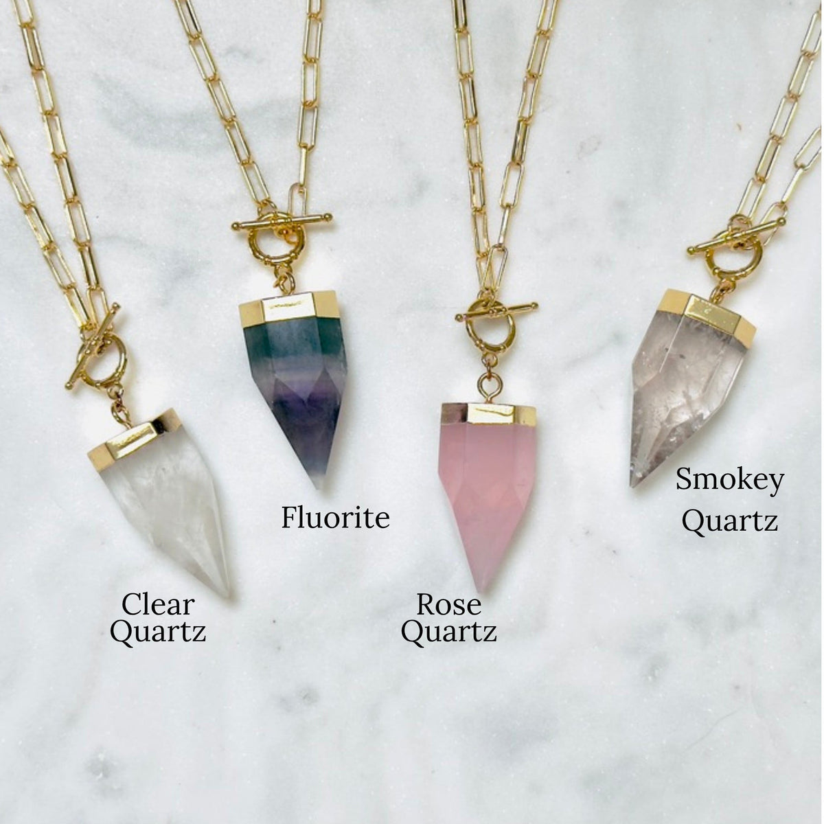 Three gold-tone chain necklaces with pointed crystal pendants are displayed on a light marble surface. The necklaces are labeled Clear Quartz, Fluorite, and Smokey Quartz, with the corresponding crystal pendants shown in white, dark green and purple, and pink.