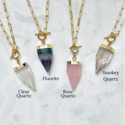 Three gold-tone chain necklaces with pointed crystal pendants are displayed on a light marble surface. The necklaces are labeled Clear Quartz, Fluorite, and Smokey Quartz, with the corresponding crystal pendants shown in white, dark green and purple, and pink.