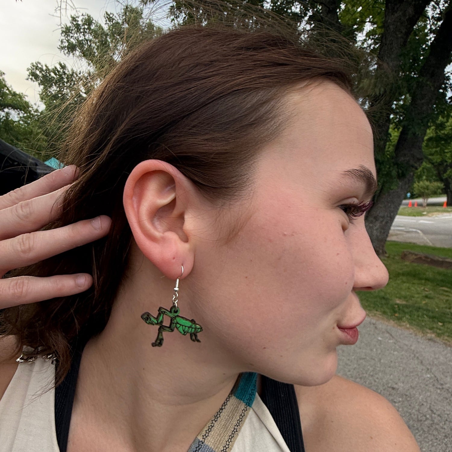 Side profile of a customer wearing hand-painted green praying mantis wooden dangle earrings. This image demonstrates the scale, movement, and eye-catching detail of these lightweight handmade earrings with hypoallergenic .925 sterling silver hooks. Ideal for insect lovers, outdoor adventurers, gardeners, and anyone looking for fun, nature-inspired wearable art.