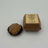 A small brown kraft paper box with a beige label sits on a plain white surface, with a round brown baked good or cookie placed below it.