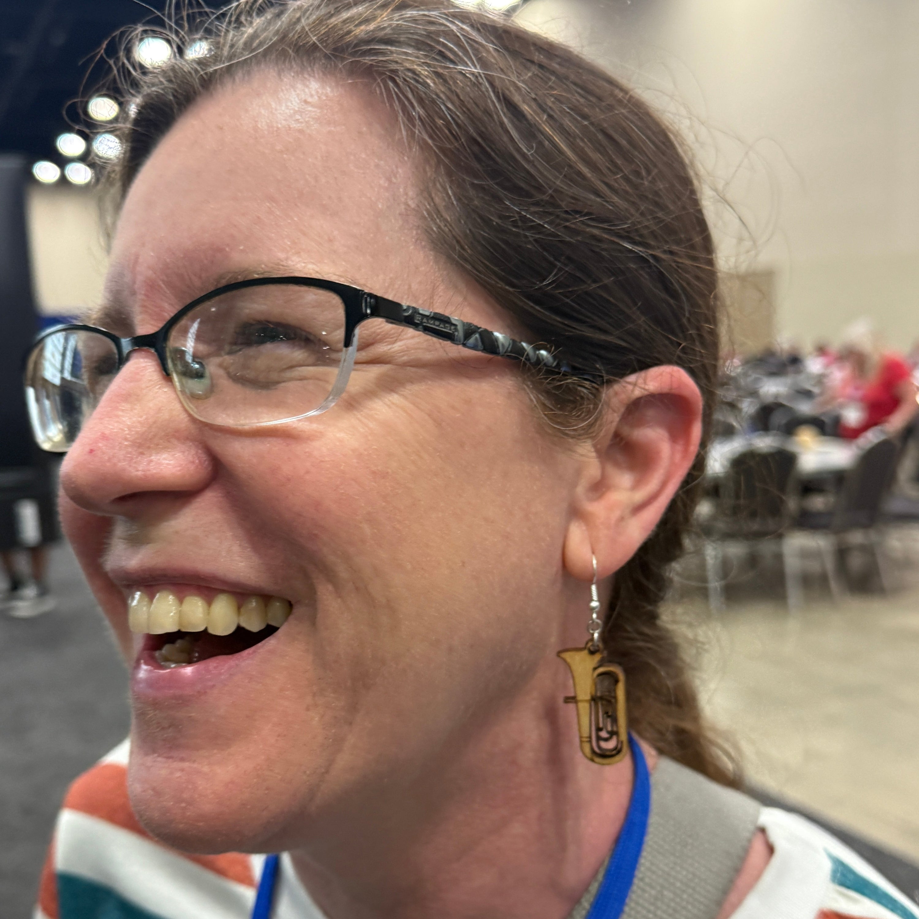 Smiling person wearing a hand-painted wooden tuba dangle earring on a hypoallergenic .925 sterling silver hook at an indoor event, demonstrating the lightweight size, natural movement, and scale of the music-themed earring when worn.