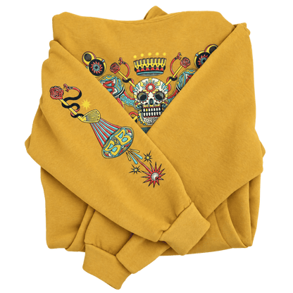 A folded mustard yellow sweatshirt featuring a colorful graphic print on the chest and sleeve. The graphic depicts a sugar skull under a crown, a snake, a teardrop, and a starburst in red, blue, black, and white.