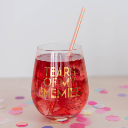"Tears of My Enemies" stemless glass with a red drink and a striped straw amid pastel confetti.