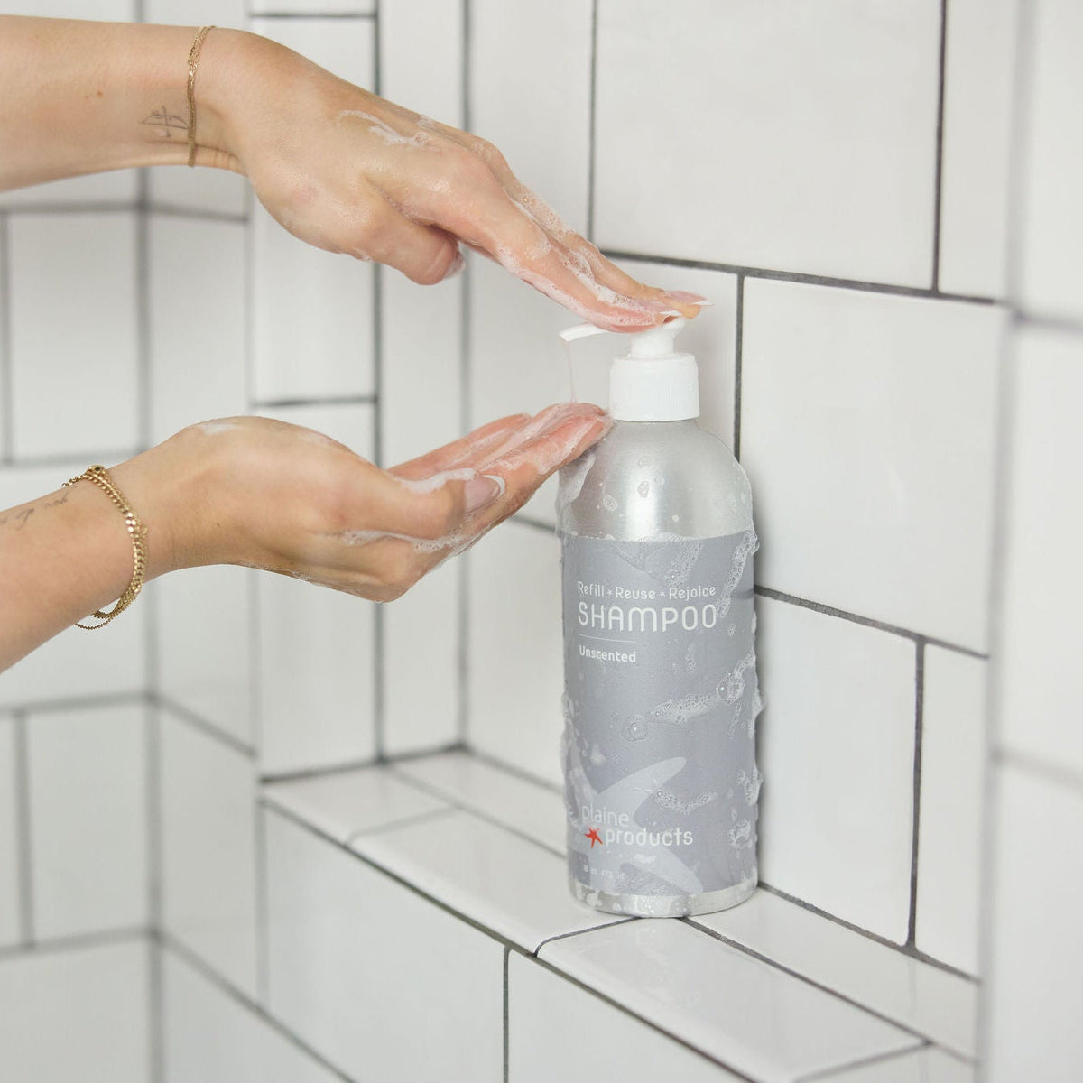 Person applying unscented, fragrance-free shampoo to hands with a plastic-free bottle of shampoo in the foreground on a tiled shower wall.