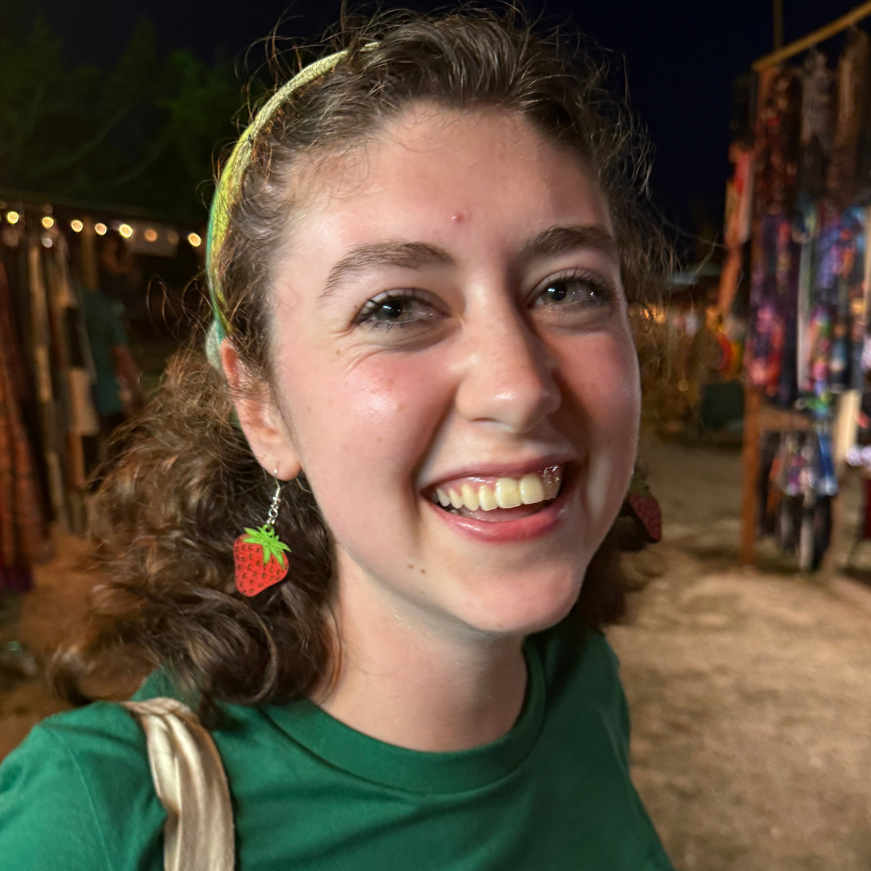 Woman wearing hand-painted wooden strawberry dangle earrings at an outdoor market, showcasing the bright red strawberry design with green leaves. This lifestyle photo highlights the size and lightweight comfort of these handmade wooden earrings with hypoallergenic .925 sterling silver hooks, perfect for summer outfits, farmers markets, fruit lovers, and everyday wear.