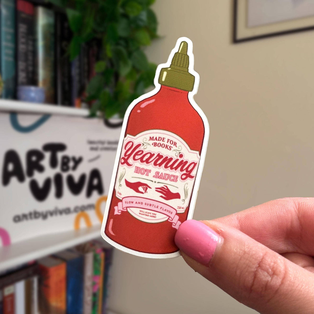 Sticker of a hot sauce bottle held by a hand in front of a bookshelf.