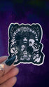 A hand with black nail polish holds a die-cut sticker featuring a black and white gothic cartoon illustration of Mickey Mouse surrounded by skulls, skeletons, and a grim reaper, set against a dark purple textured background.