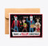 A horror-themed Christmas greeting card with a kraft paper envelope behind it, featuring illustrated characters in holiday costumes holding weapons on a black background with the text &