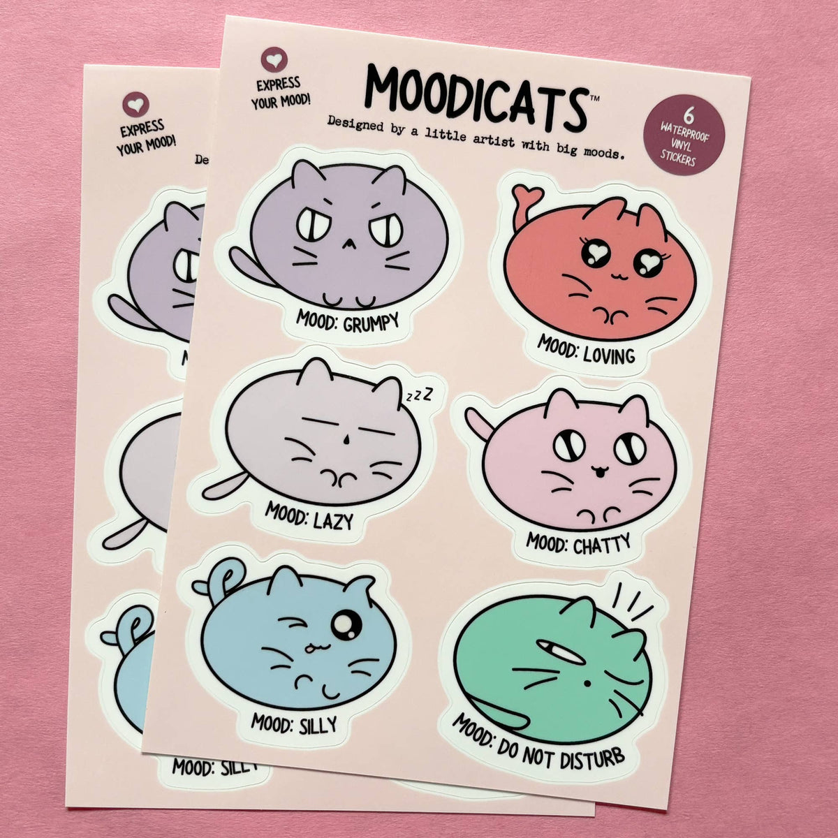 Two sticker sheets featuring cute cartoon cats in different moods, displayed on a pink background. The stickers are in pastel shades of purple, coral, gray, pink, blue, and green, with text labels like &