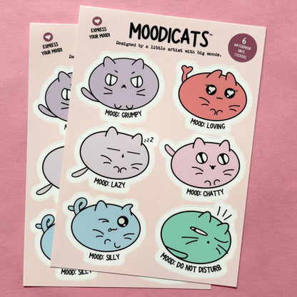 Two sticker sheets featuring cute cartoon cats in different moods, displayed on a pink background. The stickers are in pastel shades of purple, coral, gray, pink, blue, and green, with text labels like &